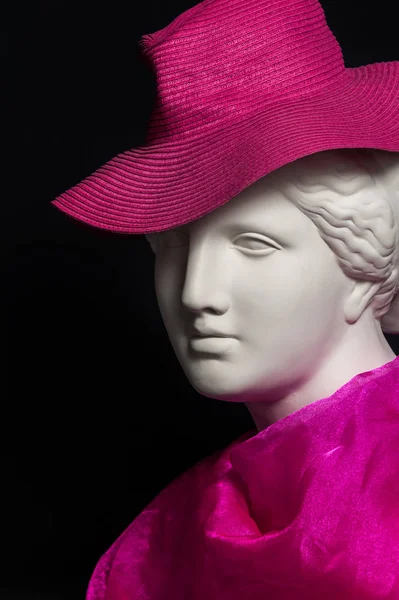 Plaster copy of ancient statue head in pink hat and scarf on a black background. Gypsum sculpture female face. — Stock Photo, Image