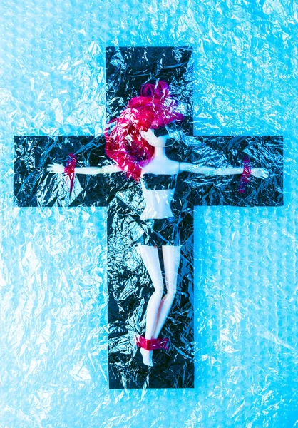 Synthetische levensduur. Kunststof pop gekruisigd op een kruis onder cellofaan film. Kunststof vervuiling en sociale problemen concept. — Stockfoto