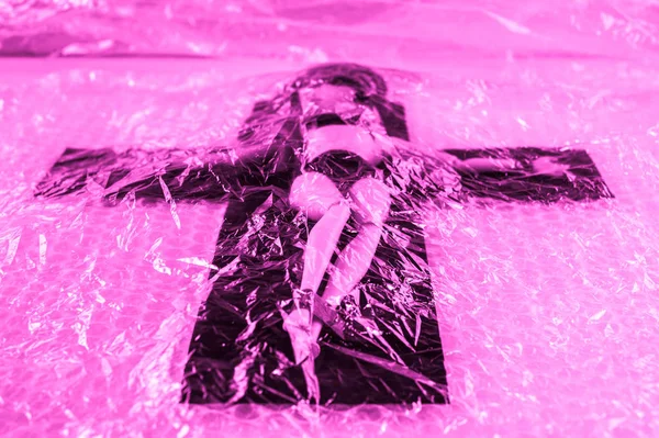 Synthetische levensduur. Kunststof pop gekruisigd op een kruis onder cellofaan film. Kunststof vervuiling en sociale problemen concept. — Stockfoto