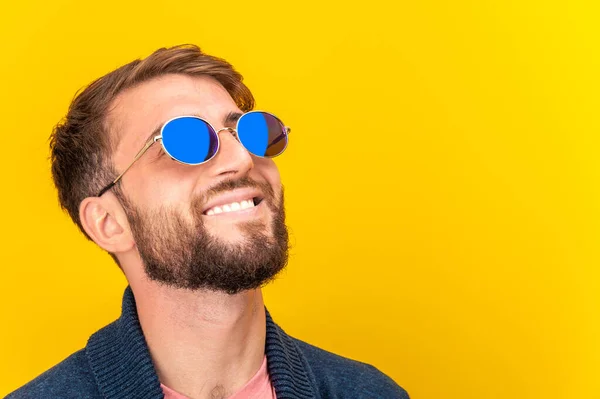 Close up portrait of funky young bearded hipster man in blue sunglasses smiling brightfully on a yellow background. Copy space. Looking up and laughing.