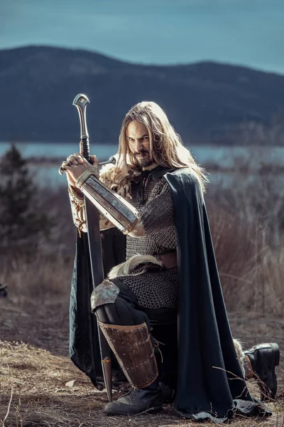 Uzun saçlı knight iki elle kılıç ile. Ortaçağ kırsal arka plan.