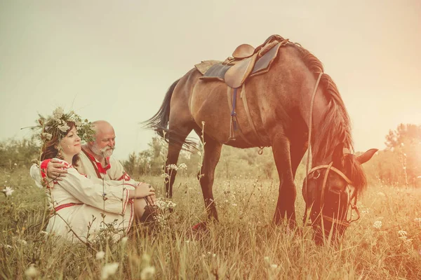 Mature man and woman in ethnic clothes are relaxing after a horse ride. Summer sunset on the background. — Stock Photo, Image