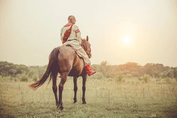 Hombre con ropa étnica está montando un caballo en el fondo de los campos de verano . —  Fotos de Stock
