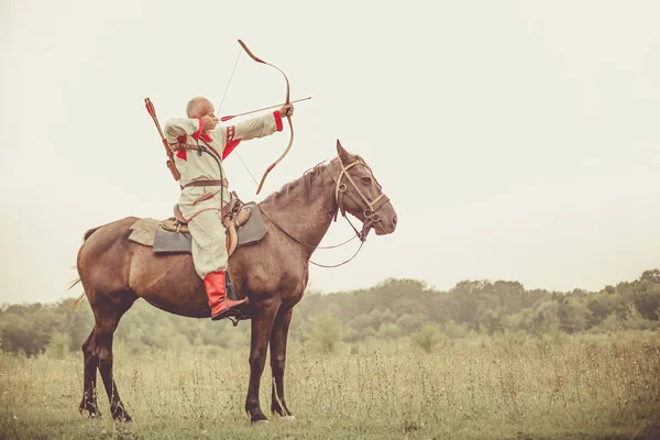 Homem de roupas étnicas está montando um cavalo e mirando a partir do arco . — Fotografia de Stock