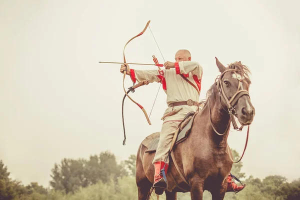 Homem de roupas étnicas está montando um cavalo e mirando a partir do arco . — Fotografia de Stock