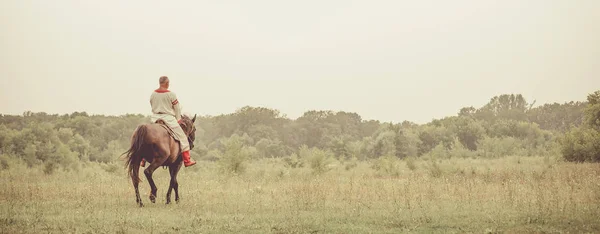 L'uomo in abiti etnici sta cavalcando un cavallo sullo sfondo campi estivi . — Foto Stock