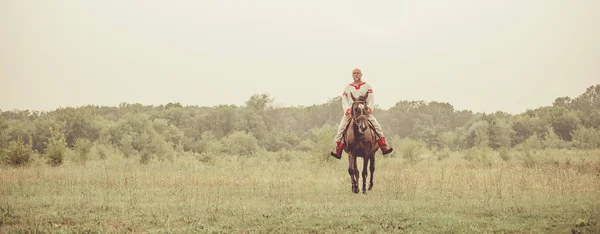 Hombre con ropa étnica está montando un caballo en el fondo de los campos de verano . —  Fotos de Stock