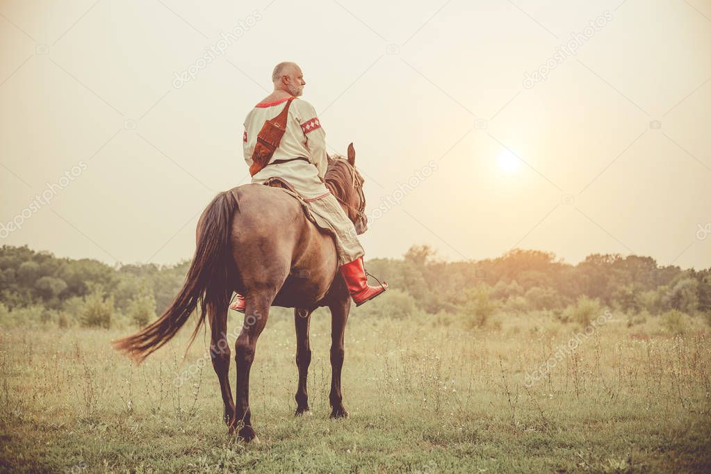 Man in ethnic clothing is riding a horse on the summer fields background.