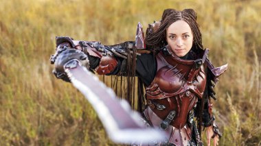 Ortaçağ şövalyesi zırhı giymiş, elinde büyük bir kılıçla günbatımı tarlalarına bakan bir kız.