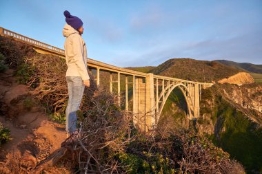 Woman tourist looking at Bixby Creek Bridge at sunset, Big Sur Area, California, USA. clipart
