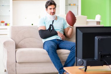 Boynu ve kolu yaralanmış bir adam televizyonda Amerikan futbolu seyrediyor.