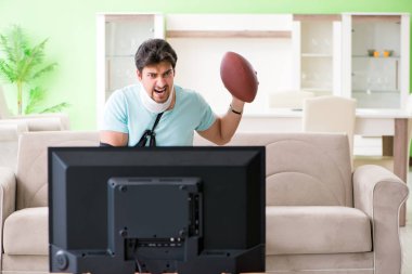 Boynu ve kolu yaralanmış bir adam televizyonda Amerikan futbolu seyrediyor.