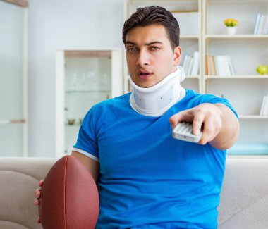 Boynu yaralanmış bir adam evde Amerikan futbolu seyrediyor.