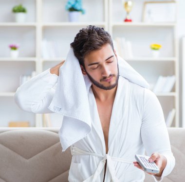 Bornozlu genç adam evde kanepede televizyon izliyor.