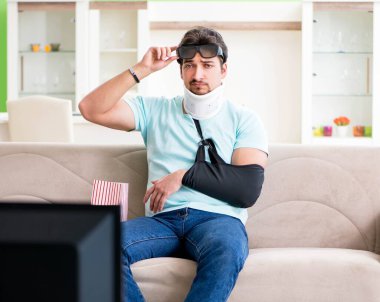 Boynu ve kolu yaralanmış bir adam televizyon izliyor.