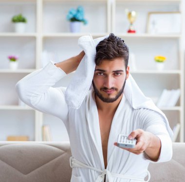 Bornozlu genç adam evde kanepede televizyon izliyor.