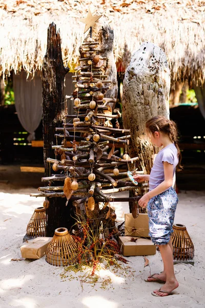小女孩正在热带旅游胜地的海滩上看那棵用木棍装饰的圣诞树 — 图库照片