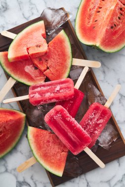 Bir yaz partisinde. Karpuz tadında ahşap bir çubuğun üzerindeki kırmızı donmuş buz, olgun karpuz parçalarıyla birlikte büyük buz parçalarının üzerinde yatıyor.                            