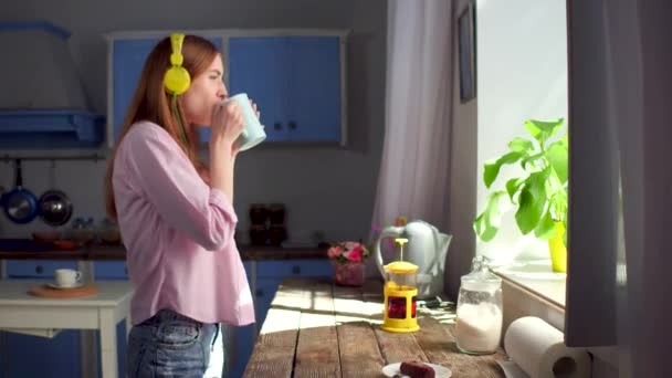 Chica en la cocina bebiendo té, escuchando música en los auriculares . — Vídeo de stock