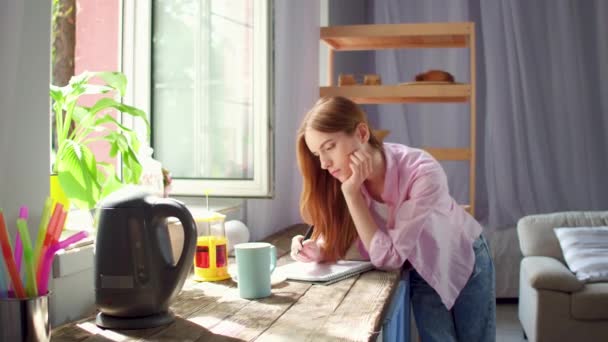 お茶を飲むと、ノートを作るキッチンの木製の机に立っている若い女性. — ストック動画