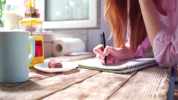 Vue rapprochée d'une jeune femme debout près d'une fenêtre ouverte prenant des notes sur un bureau en bois . — Video
