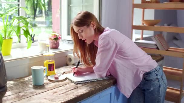 Вид на молоду дівчину, що стоїть на кухні робить нотатки . — стокове відео