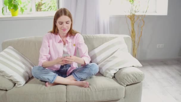 Young lady sitting on couch, typing in smartphone. — Stock Video