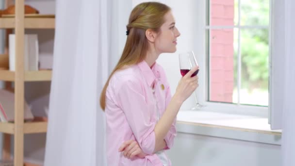 Гарненька молода жінка сидить на дивані біля відкритого вікна насолоджуючись келихом вина . — стокове відео