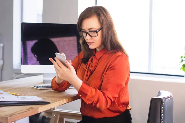 女士在办公室打字短信 穿着眼镜和深橙色衬衫的休闲女商人在她的手机上发短信 — 图库照片