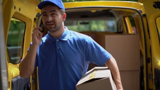 El repartidor de uniforme azul sostiene una caja de cartón mientras habla por teléfono . — Vídeos de Stock