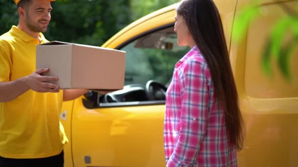 Доставщик в жёлтой униформе дает посылку клиентке возле машины — стоковое видео