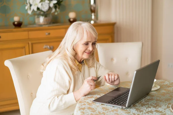 Uśmiechający się starszy kobieta używa laptopa i karty kredytowej, siedząc przy stole — Zdjęcie stockowe