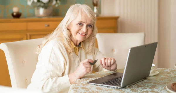 Femme âgée souriante utilise ordinateur portable et carte de crédit tout en étant assis à la table — Photo