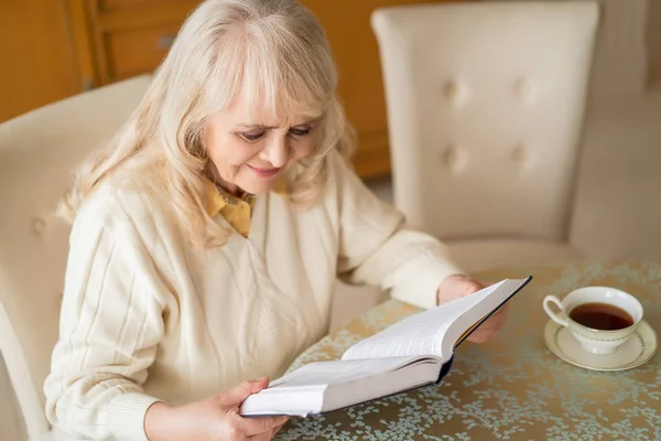 Elegante mujer mayor lee libro en la mesa y tomar el té — Foto de Stock