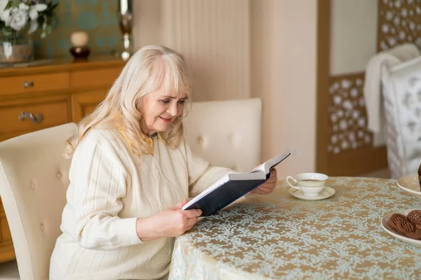 Elegant senior woman reads book at table and having tea — Stock Photo, Image