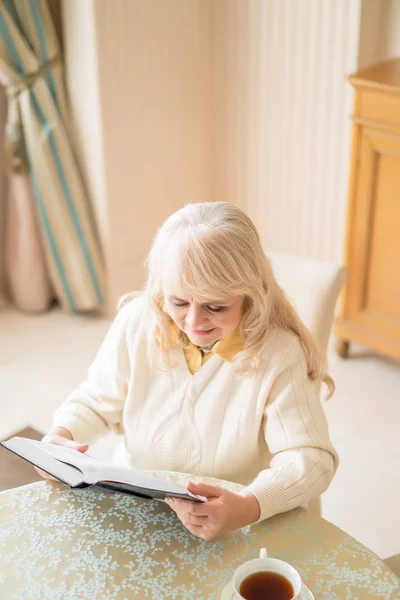 Elegante mujer mayor lee libro en la mesa y tomar el té — Foto de Stock