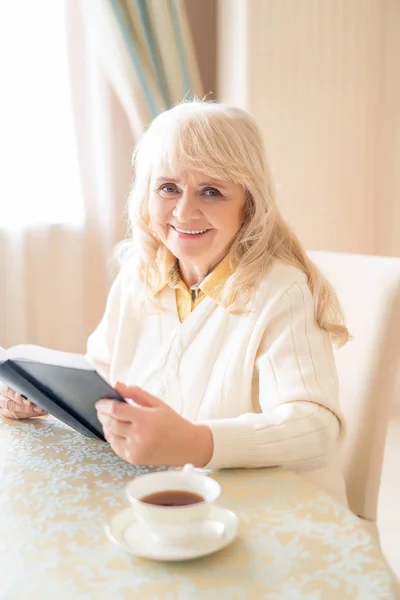 Elegante donna anziana legge il libro a tavola e prendere il tè — Foto Stock