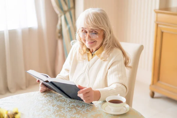 Sorridente donna anziana lettura del libro sopra una tazza di tè — Foto Stock