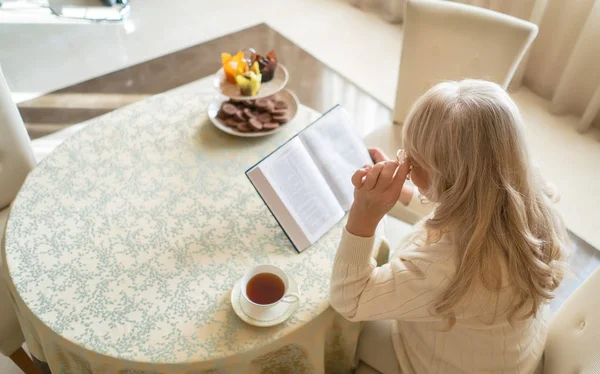 Усміхнена літня жінка читає книгу за чашкою чаю — стокове фото