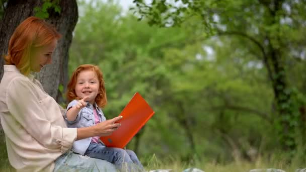 Madre y niña se sientan bajo un árbol leyendo un libro — Vídeos de Stock