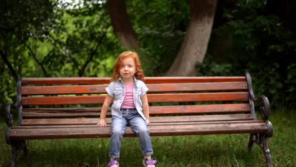 Sorrindo criança readheaded senta-se em um banco de parque . — Vídeo de Stock