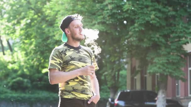 若い男は 巨大な煙を吐きます キャップとグリーンの シャツ Vaping 外側後方を着て男のクローズ アップ Vaping の明るいスローモーション撮影 — ストック動画