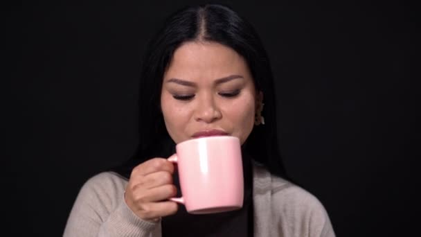 Eine brünette Frau auf schwarzem Hintergrund trinkt Kaffee mit einer rosa Tasse. — Stockvideo