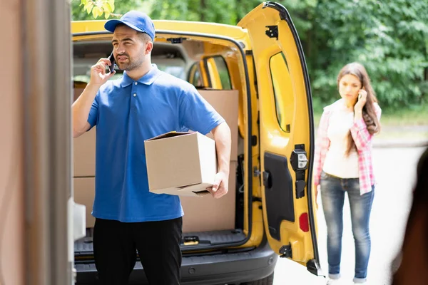 Курьер в синей форме держит картонную коробку и разговаривает по телефону. Открытый фургон в фоновом режиме . — стоковое фото