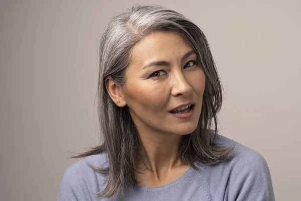 Pensive Adult Woman of Mongolian Nationality with Gray Hair on a Gray Background.