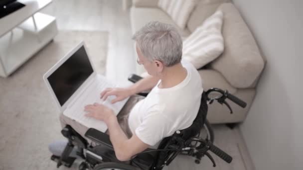 Homem de cabelos grisalhos em uma cadeira de rodas em sua ala sentado em um laptop . — Vídeo de Stock