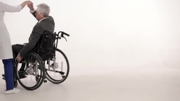 Un uomo dai capelli grigi su una sedia a rotelle sta ballando con un'infermiera dall'aspetto asiatico . — Video Stock