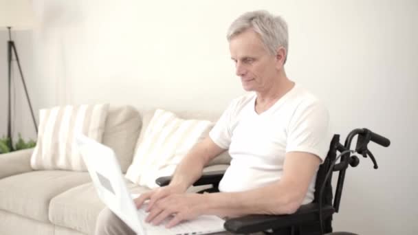 Uomo dai capelli grigi su una sedia a rotelle nel suo reparto seduto su un computer portatile . — Video Stock