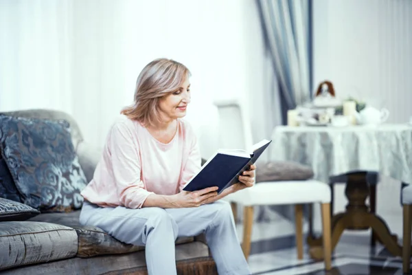 Starsza kobieta w domu ubrań siedzi na kanapie z książki w jej ręce. — Zdjęcie stockowe