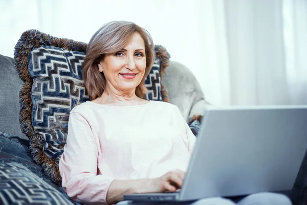 Una mujer mayor de 50 años se sienta en una computadora portátil en casa en la sala de estar . — Foto de Stock